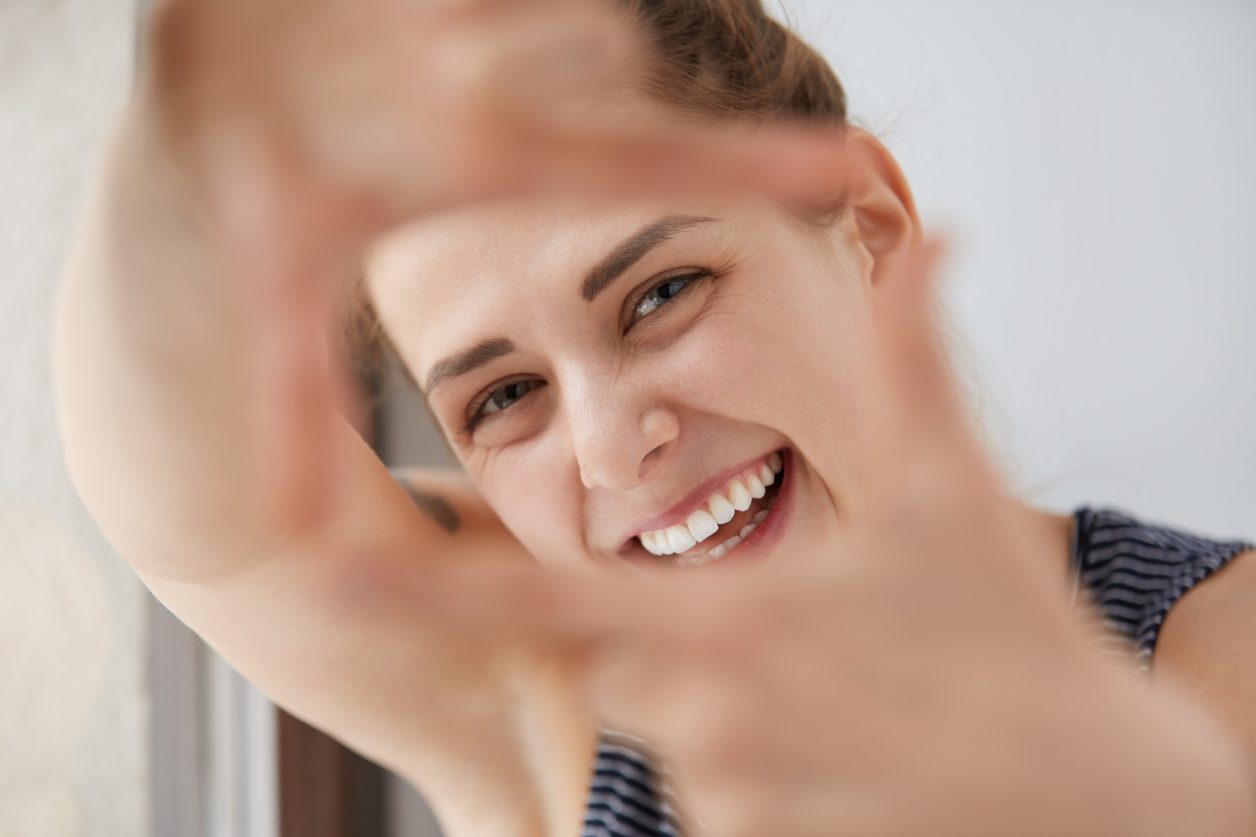 Close-up portrait of smiling lovely shiny European girl through the frame of her hands. Happy woman squinting eyes, making fun, trying to imitate photographer, being playful like little child.